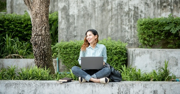 Smiling young woman sitting outdoors with a laptop, surrounded by greenery