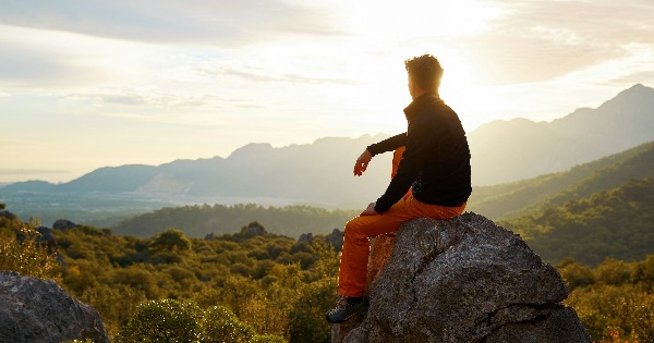 Person sitting on a mountain rock at sunset, looking over a green valley