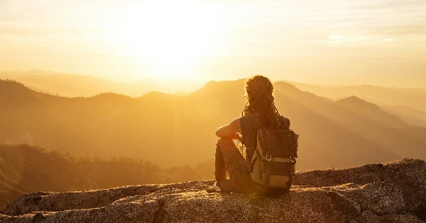 Person with a backpack sitting on a cliff edge at sunset, overlooking mountain ranges