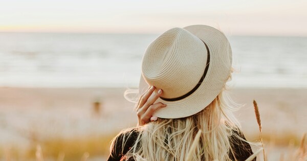 Woman in a hat sitting in golden grass at sunset, peacefully looking toward the sea