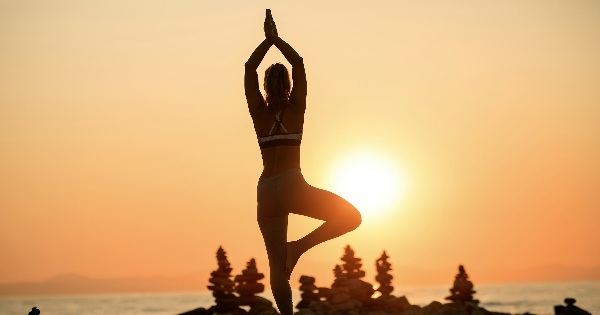 Femeie in postura yoga tree pose pe malul marii la rasarit, simbol pentru mindfulness si introspectie